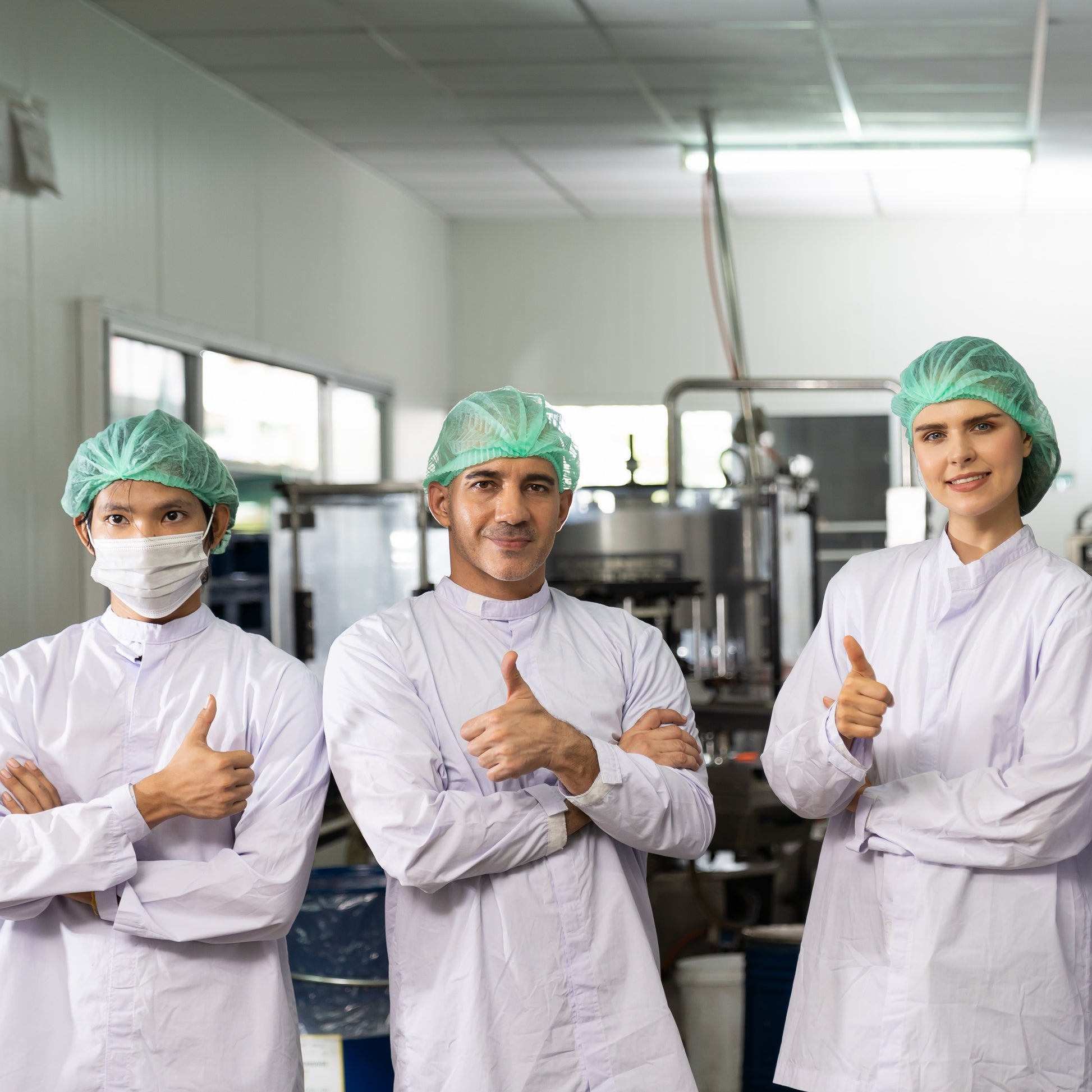 <Three smiling employees near a filling line, two male and one female, giving a thumbs up signal for wearing the correct white coat and blue hairnet. Credit> AdobeStock_454159606