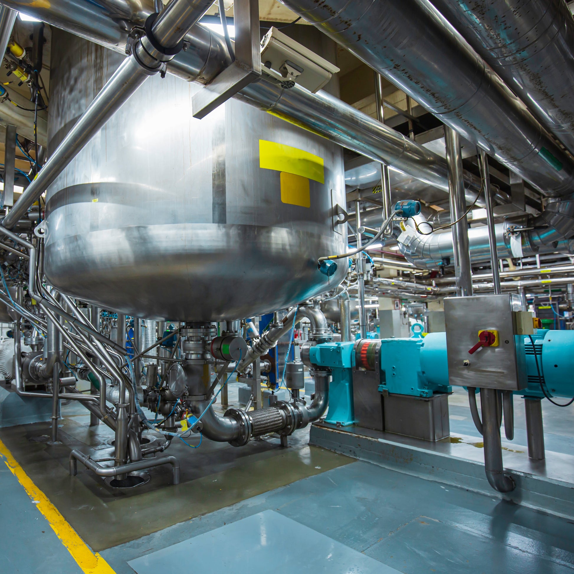 <Raised, large stainless steel mixing tank with bottom mounted homogenizer and pipework in a manufcaturing area with light blue non-slip floor. Credit> AdobeStock_467360310