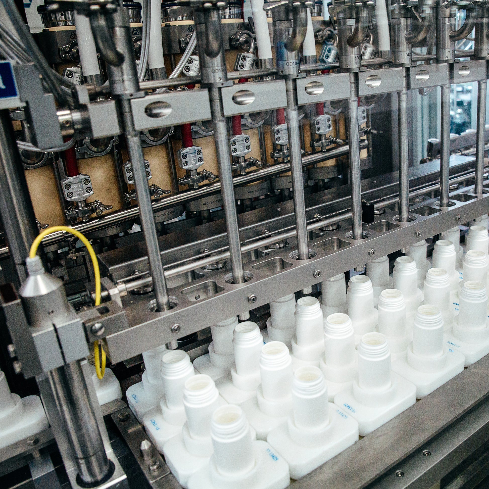 <Four rows of white bottles on a conveyor belt being filled by a series of nozzles under GMP conditions on a production line. Credit> AdobeStock_478344828