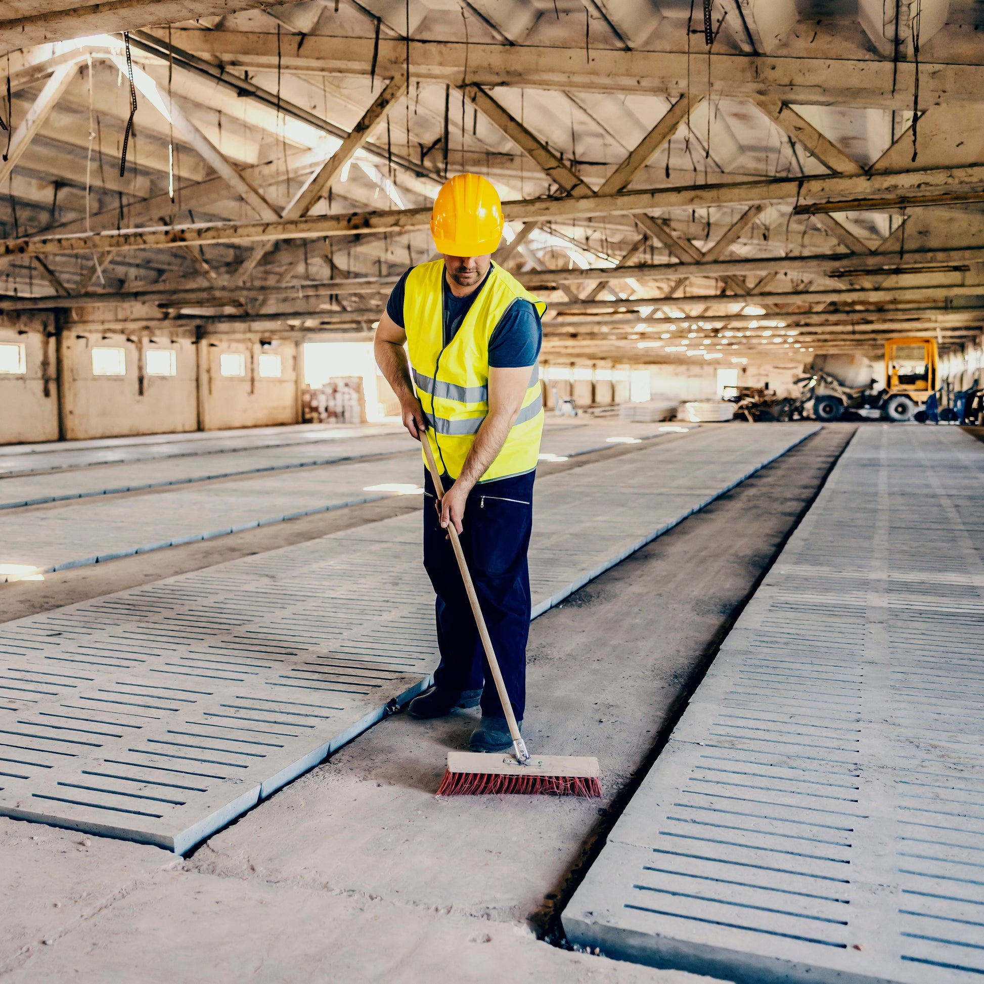 <Man wearing dark blue trousers, safety boots and a yellow fluorescent jacket and hardhat, sweeping the concrete floor of a large warehouse with a brush to remove any dust. Credit> AdobeStock_485391655