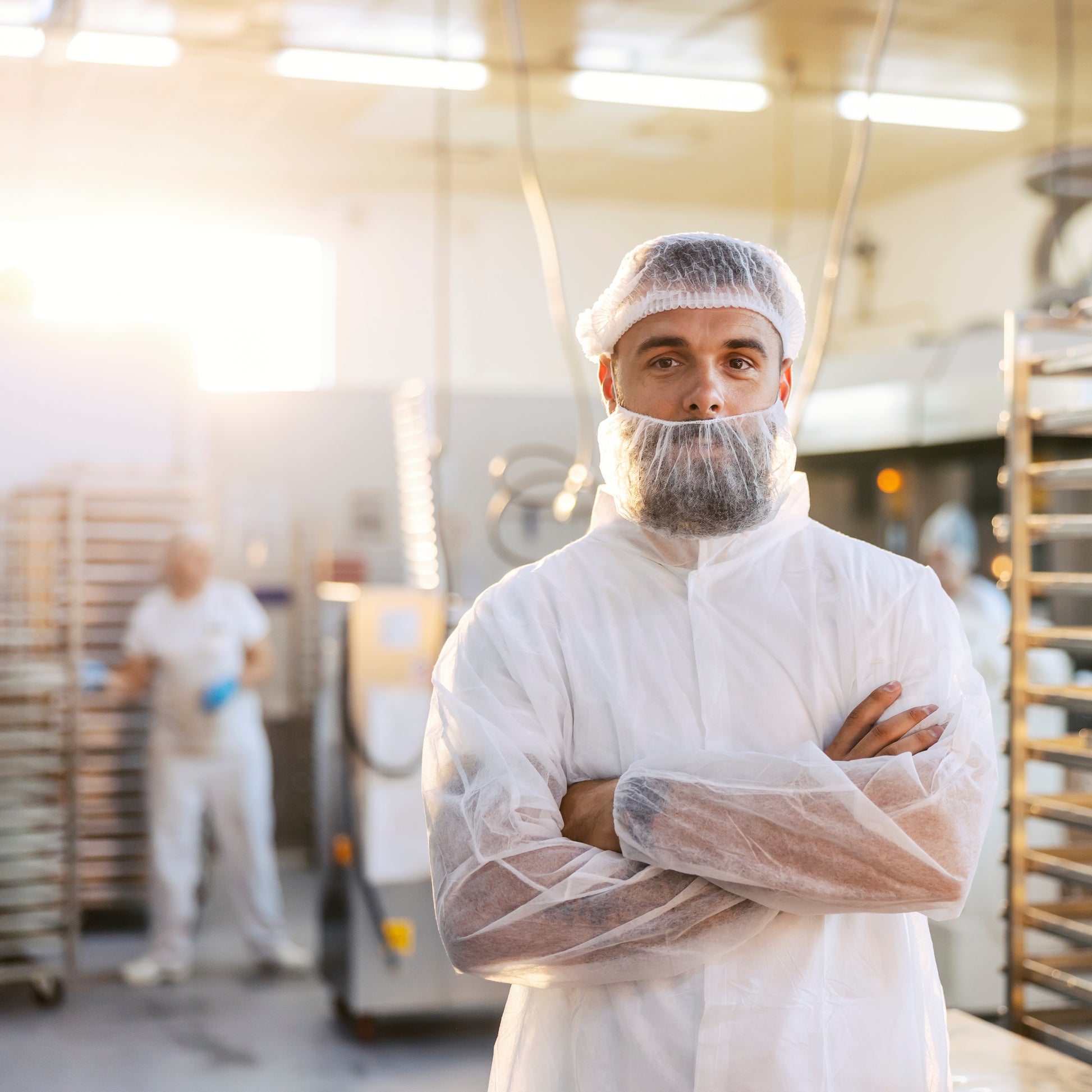 <Smiling white production operator with a moustache and crossed arms wearing a white coat, face mask and hairnet stood in a production area in front of two more similarly dressed operators in a blurred background. Credit> AdobeStock_272303046