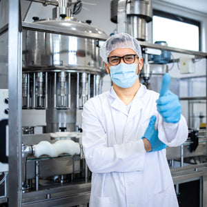 <White, smiling male production operator with glasses wearing a white coat, hairnet and face mask with blue gloves giving the thumbs up stood in front of a tablet filling machine. Credit> AdobeStock_487453879