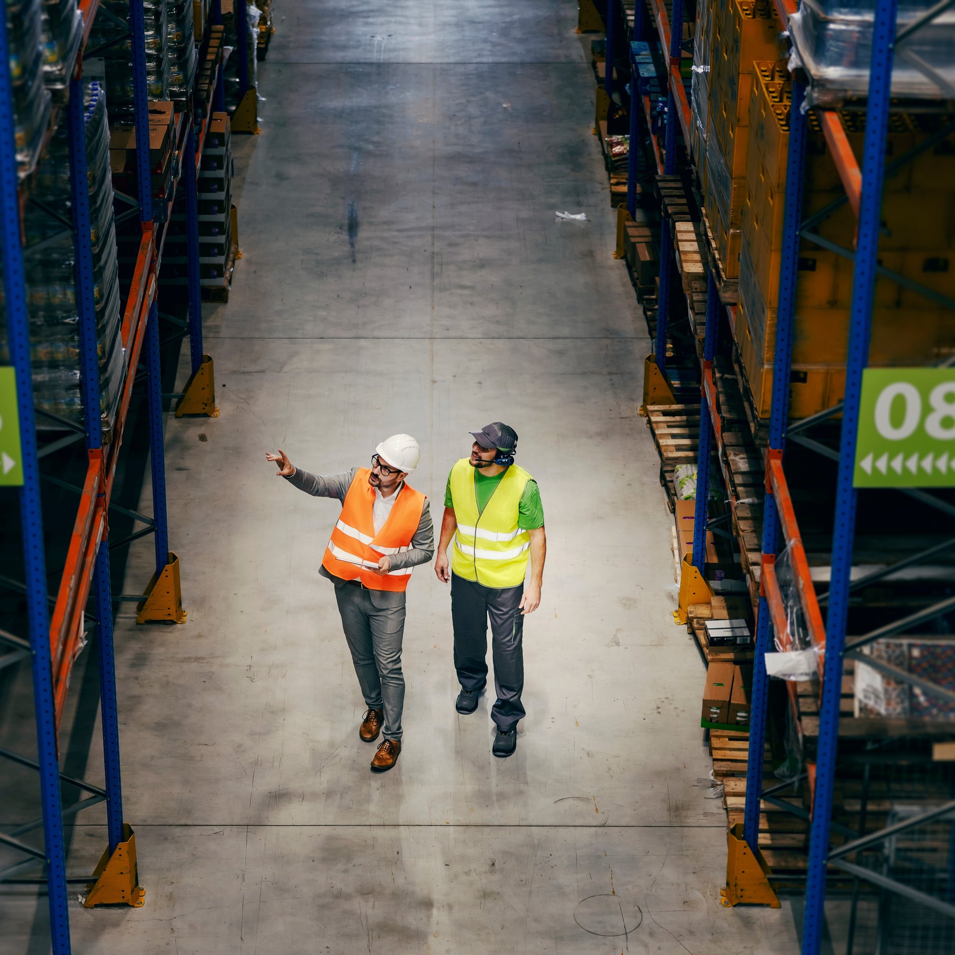 <Looking down at two male employees walking down an aisle between racking in a warehouse, one wearing orange and white hard hat pointing up, one wearing yellow and blue hard hard looking. Credit> AdobeStock_489525726