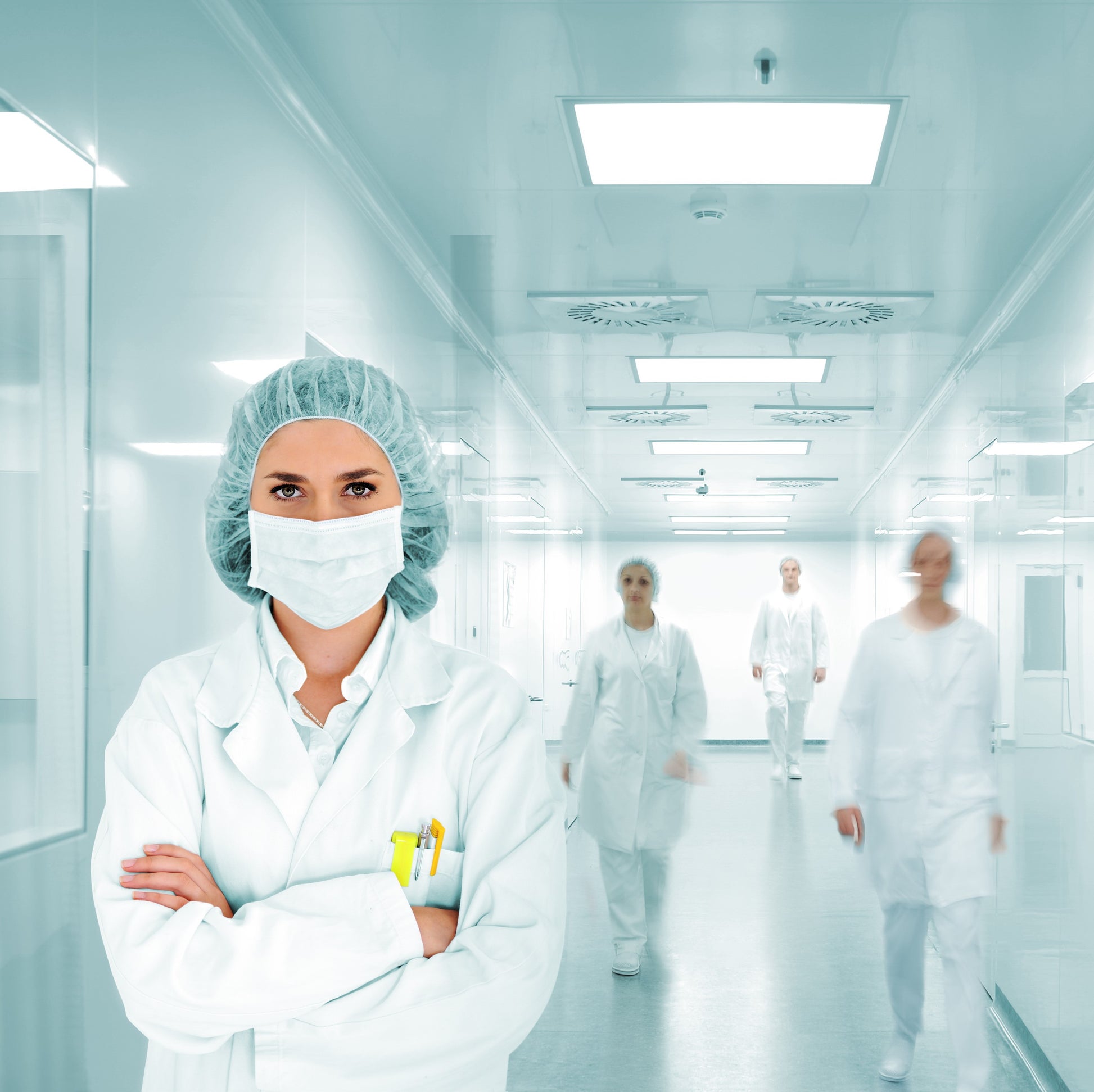 <Four female employees at various distances away walking down a white corridor in production with a series of square ceiling LED lights, windows, and non slip floor, wearing white overalls and blue hairnets. Credit> AdobeStock_49725046