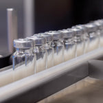 <Row of glass vaccine vials with metal caps passing down a conveyor belt with guardrails keeping them in place to be filled. Credit> AdobeStock_513373066