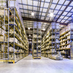 <Clean, well lit, large warehouse with concrete floor, racking containing pallets of boxes of materials in rows seven high and twenty wide, with white sloping ceiling with black metal square frame. Credit> AdobeStock_51465184