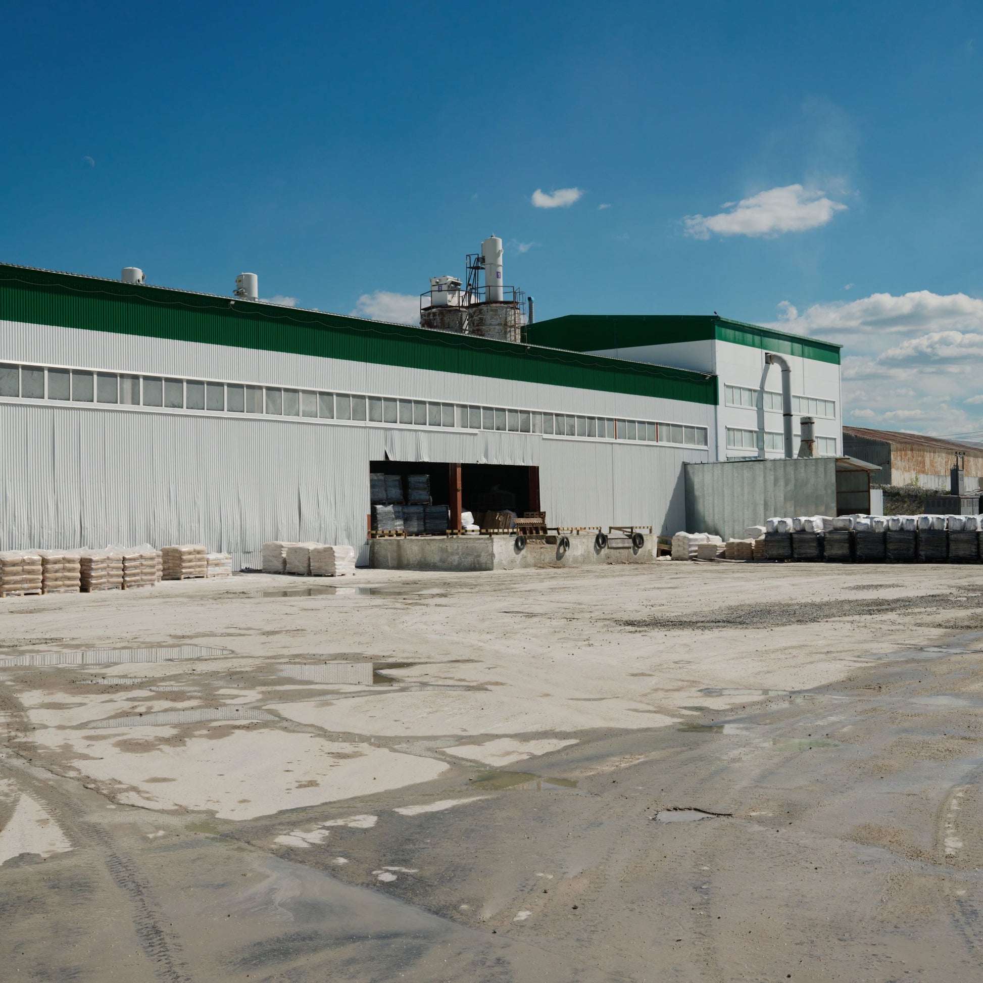 <Concrete yard, in sunshine after rain, of a grey and green striped corrugated metal dietary supplement production  building taking in pallet deliveries of materials through a shutter door. Credit> AdobeStock_525171795