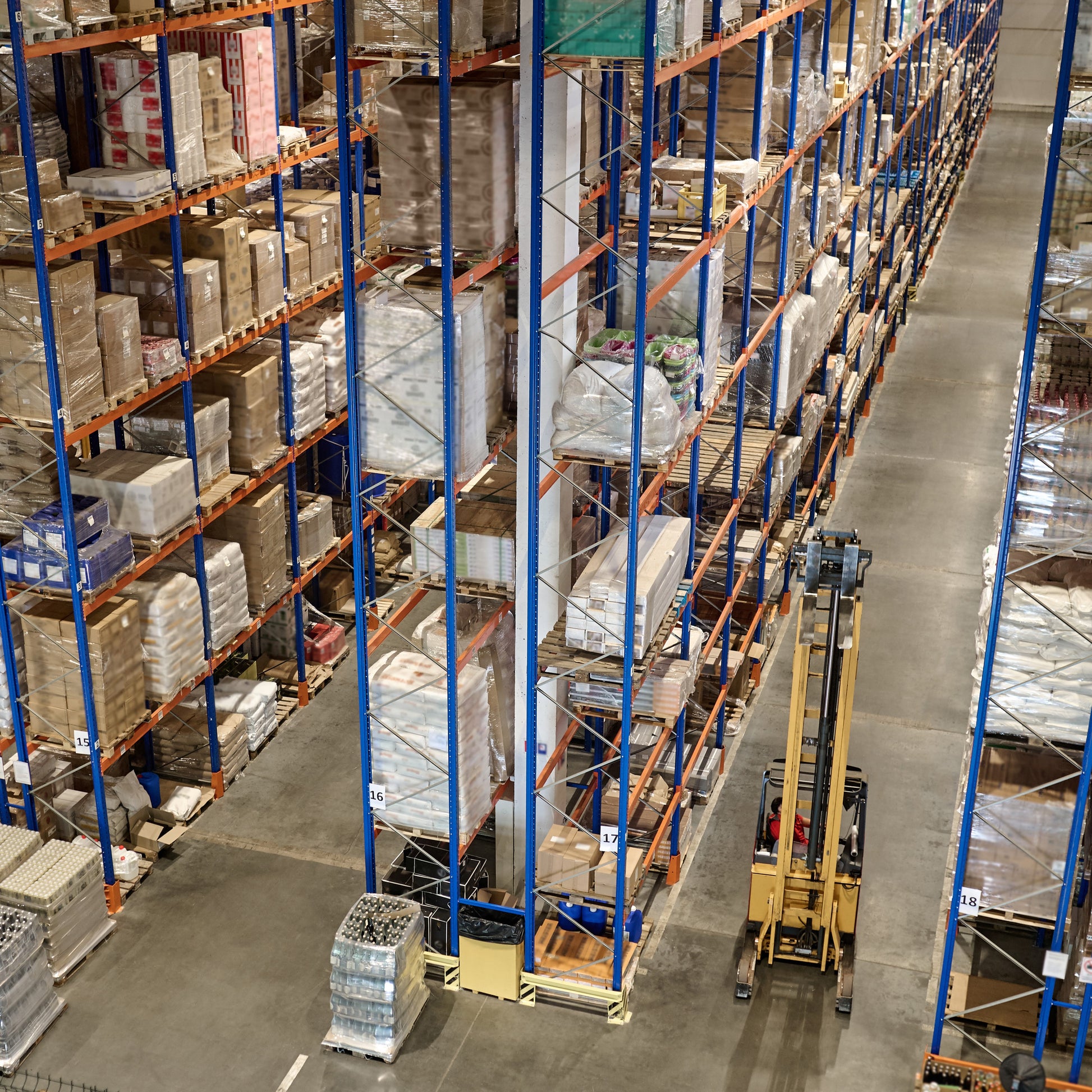 <View from the ceiling looking down into clean, tidy warehouse to see a yellow forklift truck on concrete floor operating between rows of racking containing boxes and sacks of materials. Credit> AdobeStock_544570194