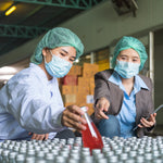 <A female Quality Assurance Officer reviewing dietary supplement bottles filled with red liquid with a female Production Team Leader, both wearing blue hairnets and face masks. Credit> AdobeStock_551742643