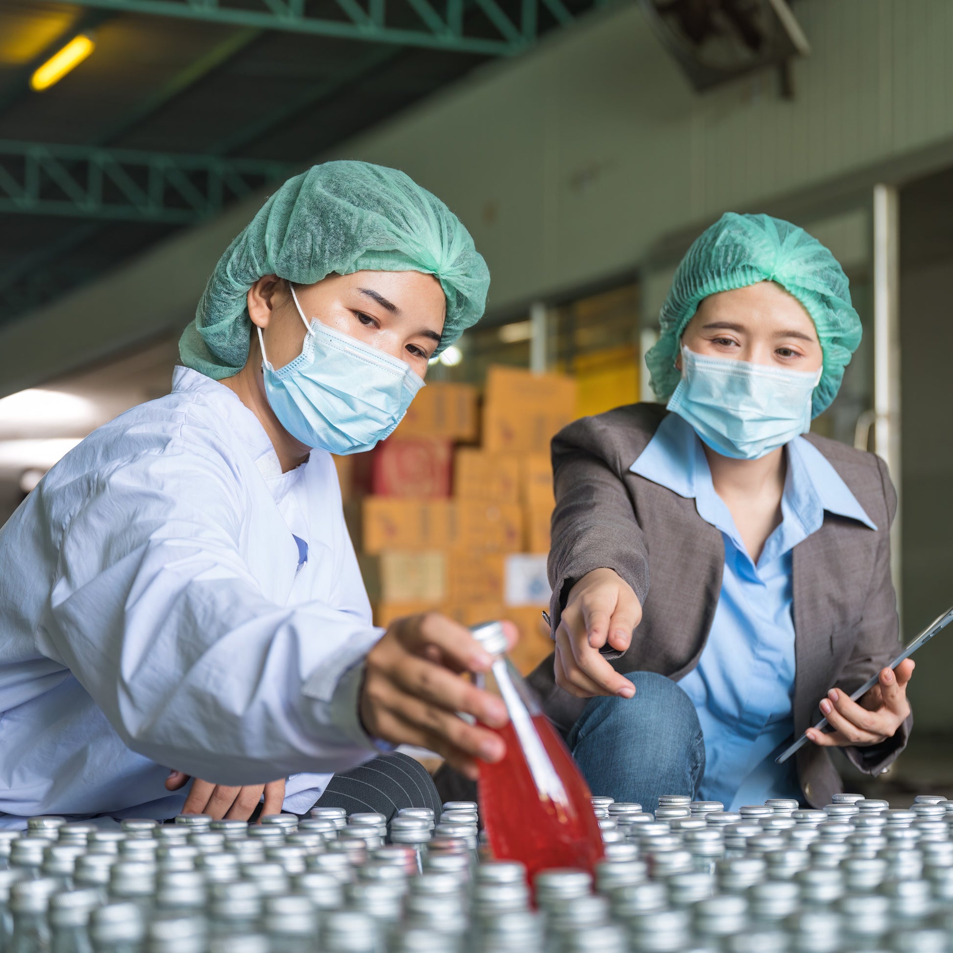 <Female QA officer pointing to a specific sample in the middle of hundreds of bottles with silver metal caps, with a female Production operator picking it up revealing a dark red liquid inside. Credit> AdobeStock_551742643