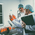 <Two Quality Officers, one male with a clipboard, the other female with a laptop, both wearing white coats and blue hairnets, inspecting bottles of orange liquid running down a production line conveyor belt,. Credit> AdobeStock_552543492