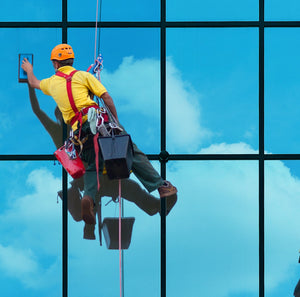 <Male window cleaner with orange hard hat and yellow T-shirt  abseiling down the face of a glass building cleaning each window from a bucket and sponge. Credit> AdobeStock_561040865