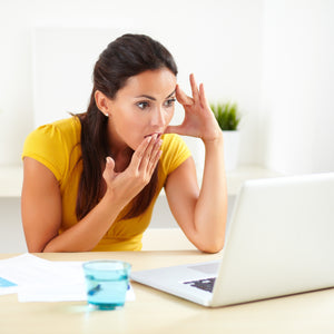 <Woman wearing yellow top sitting at a table, with white background, looking shocked holding her hand to her mouth at an e-mail response to a complaint on a white laptop. Credit> AdobeStock_78493419