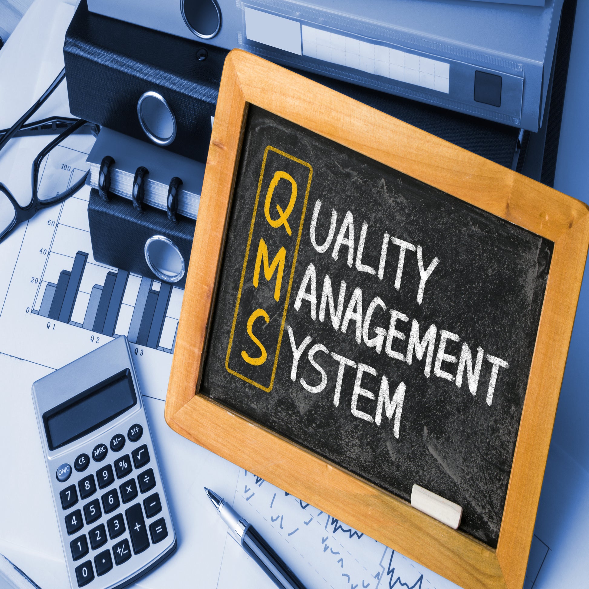 <Small, wooden framed blackboard with Quality Management System written in white chalk, except for the letters QMS written and circled in yellow, sitting on a desk against some box files next to a calculator, pen and glasses. Credit> AdobeStock_84985243