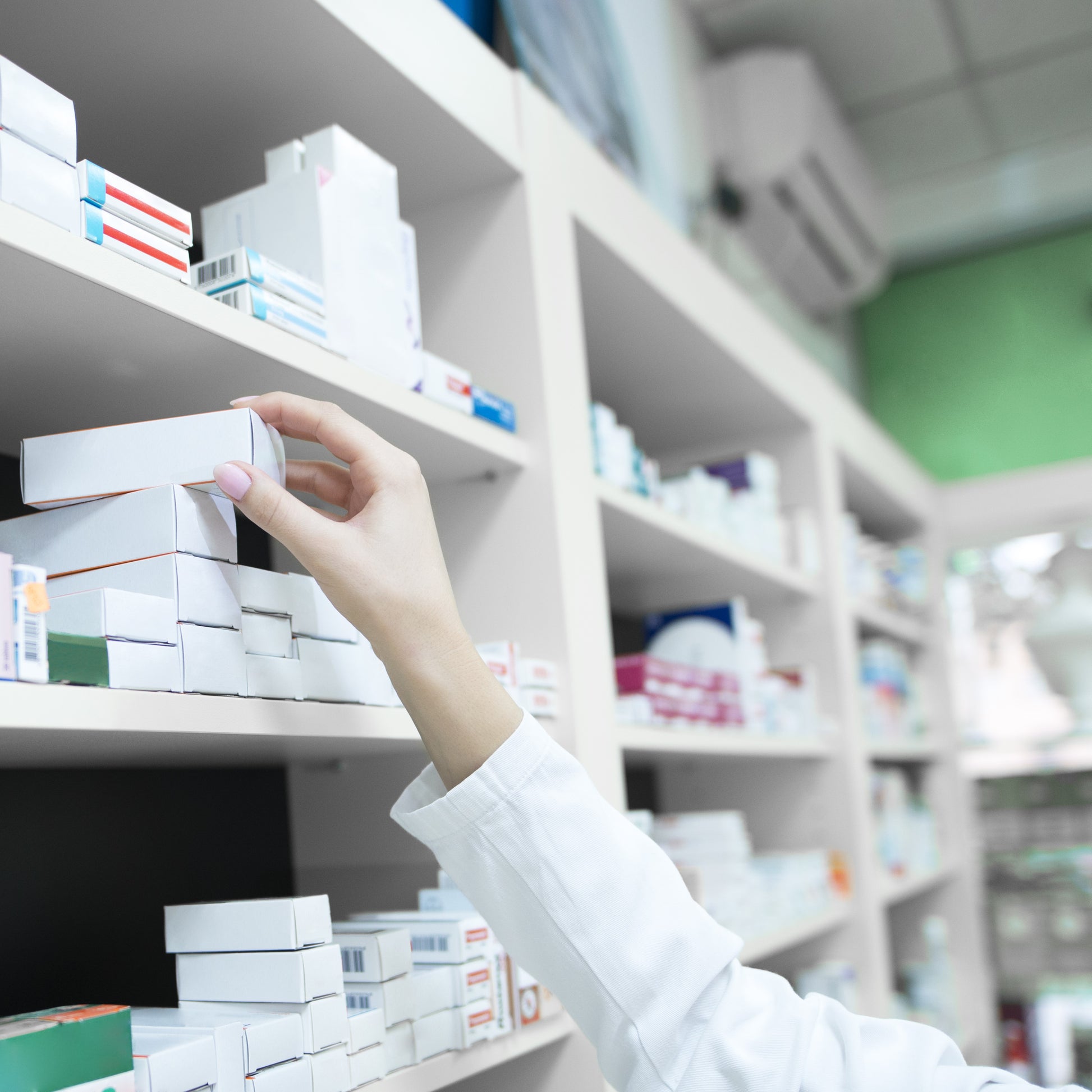 <Close up of female pharmacist arm selecting a cream tube in a carton from the shelf of medicines behind the counter. Credit> Aleksandarlittlewolf @ Freepik