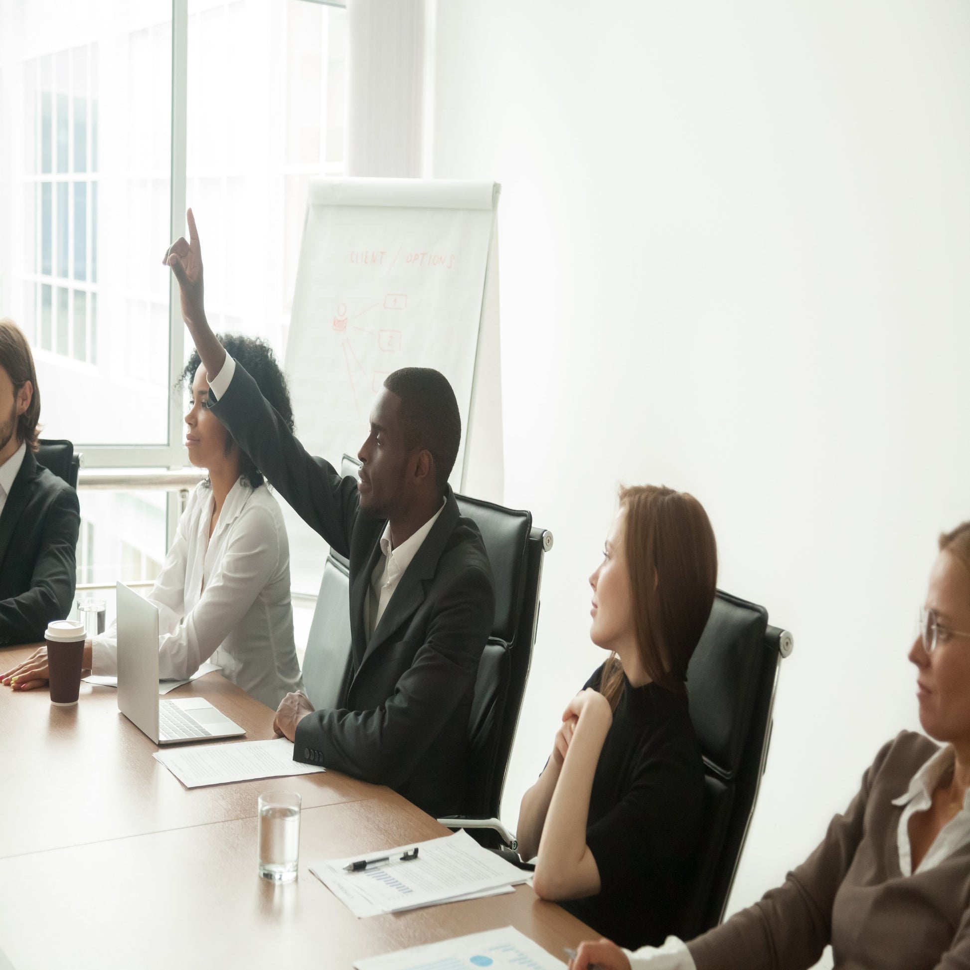 <Five employees, three female, two male, in a training session in a white meeting room with wooden desk and flipchart, one of the males holding his hand up to ask a question. Credit> 116558699 © Fizkes | Dreamstime.com