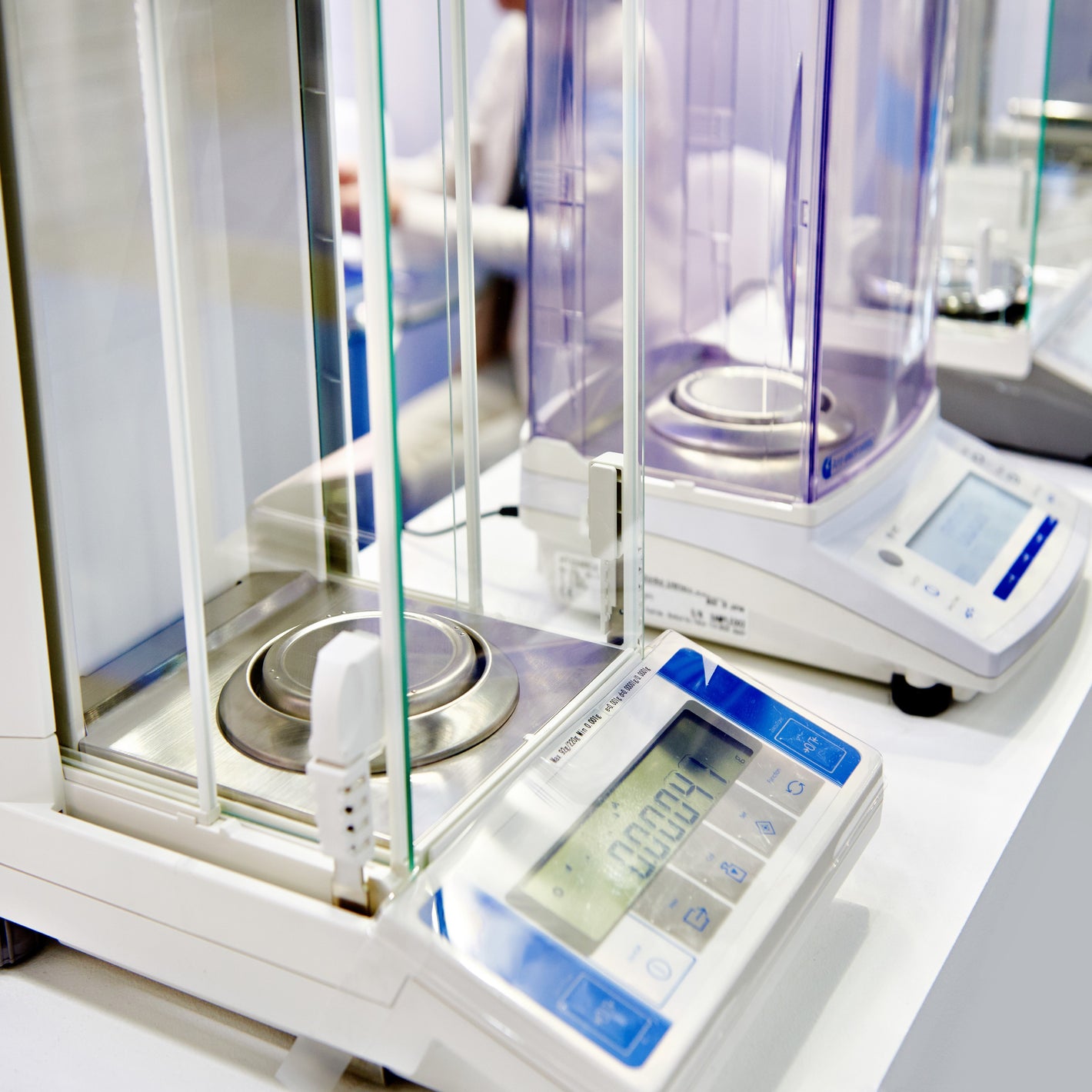 <Row of four digital analytical balances with round stainless steel weigh plate and glass or plastic screens on a white laboratory worktop with female analyst working behind. Credit> 147005408
© Ryzhov Sergey | Dreamstime.com