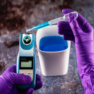 <Analyst wearing purple gloves pipetting a sample of clear, blue liquid from a white beaker on a stone effect worktop onto the circular prism of a handheld refractometer. Credit> 210828898 © Powerofflowers | Dreamstime.com