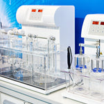 <Automated dissolution tester, a tall white unit with red digital display of time, three large glass beakers with metal apparatus inside placed in a  plastic tray, all sat on a white lab surface next to a disintegration tester, with a bright blue wall behind. Credit>  219341697 © Ryzhov Sergey | Dreamstime.com