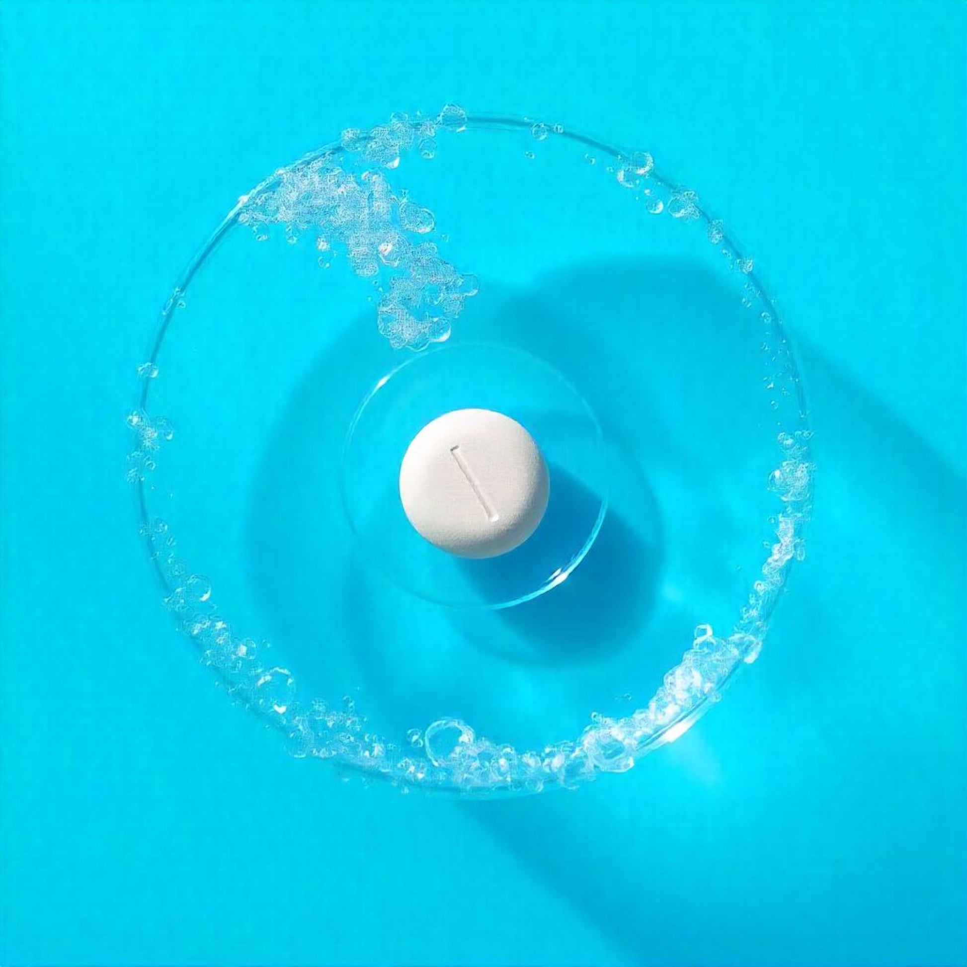 <Overhead view of a round white tablet, not showing any signs of breaking down, in a glass of warm water sat on a bright light blue work surface. Credit> Freepik