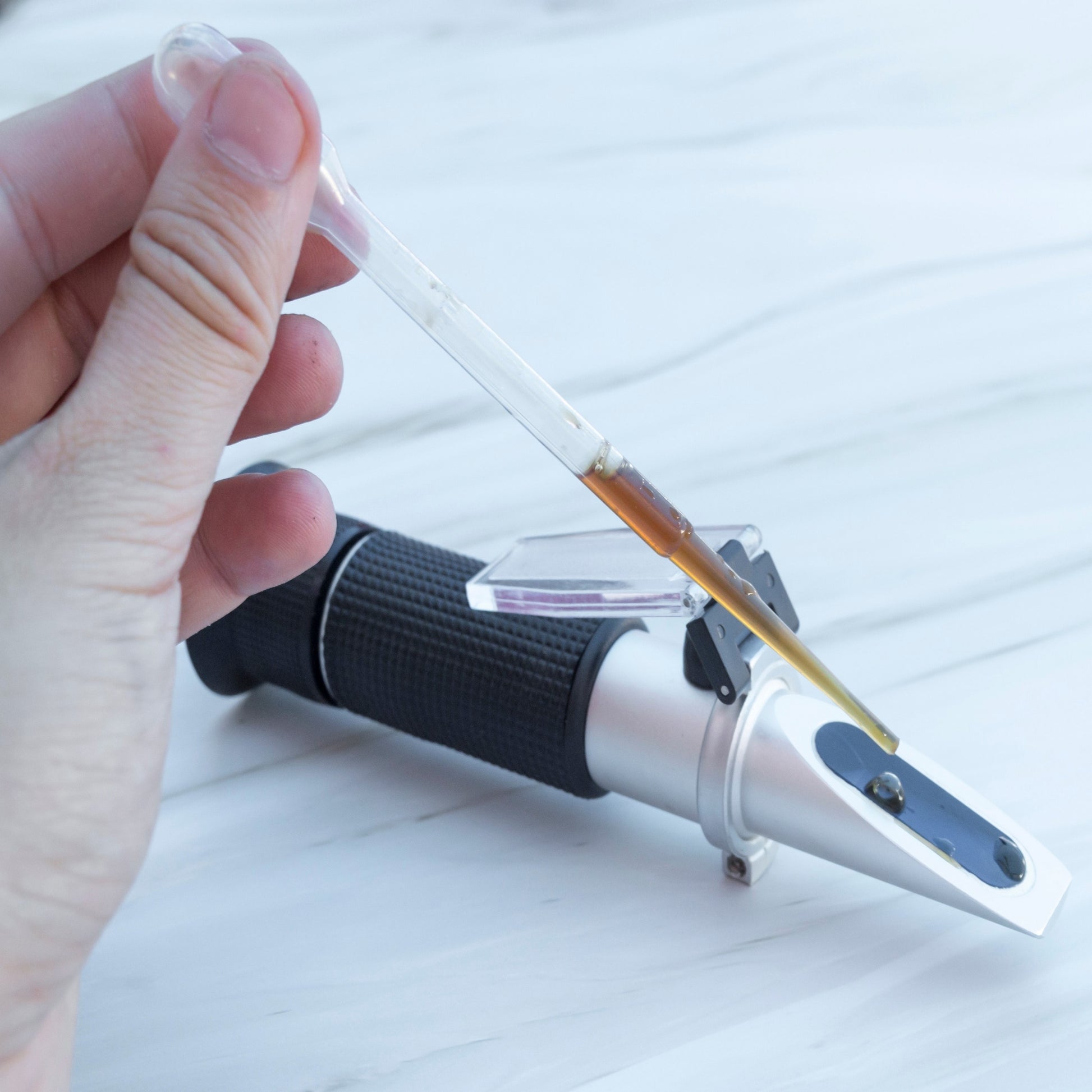 <Analyst pipetting a brown liquid onto the triangular shaped prism of a handheld refractometer on a white stone effect worktop. Credit> 2173352003 by Svarun @ Shutterstock